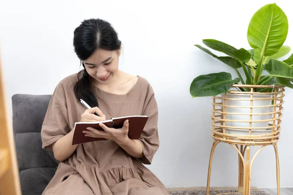 Asian woman sits in her living room writing a diary or notebook on the carpet, Weekends or holidays at home, Relaxation, Happy time, Living at home, Sit comfortably on vacation, Comfortable corner.