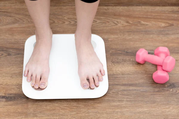 Woman\'s feet stepping on electronic scales, Measure your body weight after a workout or yoga session, Weight control, Health care, Dumbbell exercise, Background is light brown wood grain.