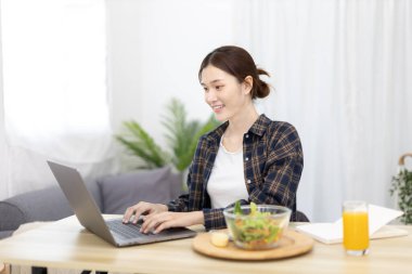 Beautiful woman using on a laptop on a table with vegetable salad and freshly squeezed orange juice for breakfast, Morning break on weekends, Comfort zone, Happy relaxation,  Stay home.