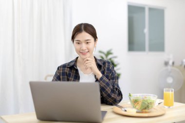 Beautiful woman using on a laptop on a table with vegetable salad and freshly squeezed orange juice for breakfast, Morning break on weekends, Comfort zone, Happy relaxation,  Stay home.