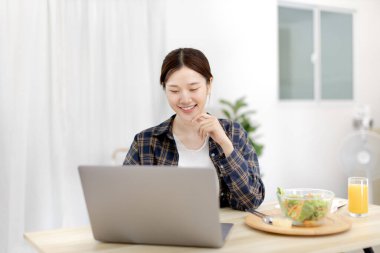Beautiful woman using on a laptop on a table with vegetable salad and freshly squeezed orange juice for breakfast, Morning break on weekends, Comfort zone, Happy relaxation,  Stay home.