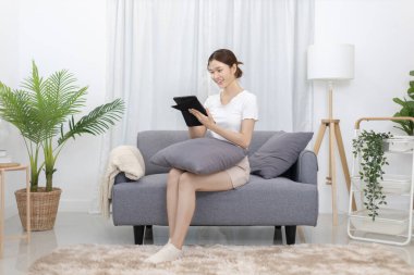 Asian woman sitting on the sofa with a tablet, Looking at tablet screen, Relaxing at home, Relaxing at home, Weekend activities, Comfort Zone, Happy working lifestyle, Happy lifestyle, Feel good.
