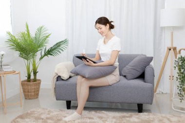 Asian woman sitting on the sofa with a tablet, Looking at tablet screen, Relaxing at home, Relaxing at home, Weekend activities, Comfort Zone, Happy working lifestyle, Happy lifestyle, Feel good.