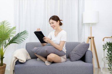 Asian woman sitting on the sofa with a tablet, Looking at tablet screen, Relaxing at home, Relaxing at home, Weekend activities, Comfort Zone, Happy working lifestyle, Happy lifestyle, Feel good.