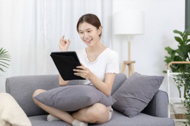 Asian woman sitting on the sofa with a tablet, Looking at tablet screen, Relaxing at home, Relaxing at home, Weekend activities, Comfort Zone, Happy working lifestyle, Happy lifestyle, Feel good.