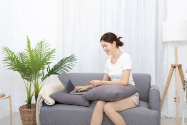 Asyalı bir kadın kanepeye uzanıyor ve kredi kartıyla online alışveriş yaparak ödeme veya çevrimiçi işlemler, mali işlemler ve internet güvenliği için kredi kartı alışverişi yapıyor..
