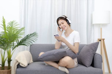 Beautiful Asian woman happily wearing headphones listening to music wirelessly in smart phone, Artist mood, Dance or move to the beat of the music, Listening to music on the sofa in the living room.
