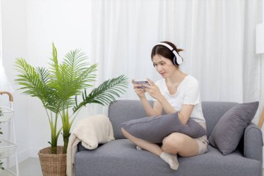 Beautiful Asian women are playing games on their mobile phones or smartphones with pleasure and fun, Put on headphones to play games, Girl's favorite corner on weekends,  Happy living room.