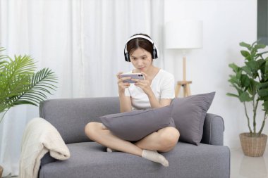 Beautiful Asian women are playing games on their mobile phones or smartphones with pleasure and fun, Put on headphones to play games, Girl's favorite corner on weekends,  Happy living room.