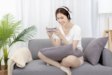 Beautiful Asian women are playing games on their mobile phones or smartphones with pleasure and fun, Put on headphones to play games, Girl's favorite corner on weekends,  Happy living room.