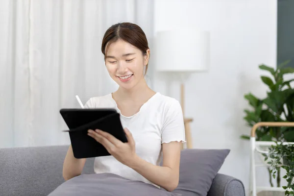 Asian woman sitting on the sofa with a tablet, Looking at tablet screen, Relaxing at home, Relaxing at home, Weekend activities, Comfort Zone, Happy working lifestyle, Happy lifestyle, Feel good.