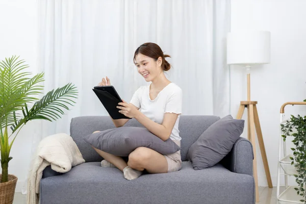 Asian woman sitting on the sofa with a tablet, Looking at tablet screen, Relaxing at home, Relaxing at home, Weekend activities, Comfort Zone, Happy working lifestyle, Happy lifestyle, Feel good.