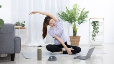 Spor kıyafetleri içinde güzel bir Asyalı kadın ısınıyor ya da evde dambıllar, protein içeceği ya da bir şişe su ile hafif egzersizler yapıyor ve evde dizüstü bilgisayar kullanıyor..