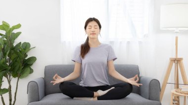 Spor kıyafetlerindeki güzel genç kadın meditasyon yapıyor ya da yoga yapıyor ve kanepede nefes alıyor, vücut ve zihin sağlığı, meditasyon yoluyla rahatlama, sağlıklı egzersiz, sessiz oturma odası....