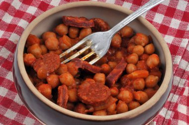 Bir kase nohut ve çatallı sucuk kareli peçeteye yakın duruyor. 