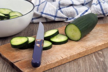 Sliced cucumber with a knife on a cutting board with a salad bowl of cucumber slices