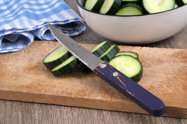 Sliced cucumber with a knife on a cutting board with a salad bowl of cucumber slices