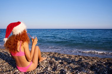 Noel Baba şapkası takan güzel kızıl saçlı kadın ve pembe bikinili okyanus kıyısında kış tatilinin tadını çıkarıyor. Noel ya da Noel tatilleri deniz kenarında lüks bir tatil köyünde. Gezgin kız kokkuyruk içiyor.
