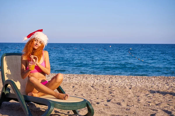 美丽的红头发女人头戴圣诞帽 身穿粉色比基尼 在海边享受寒假 圣诞节或新年假期在海滨的豪华度假胜地 旅行女孩喝燕尾服 — 图库照片