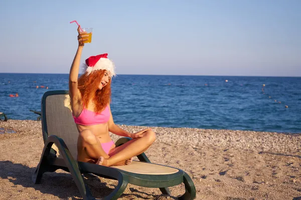 サンタの帽子とピンクのビキニを身に着けている美しい赤い髪の女性は 海の近くのビーチで冬の休暇を楽しんでいます 旅行者の女の子はカクテルを飲む — ストック写真