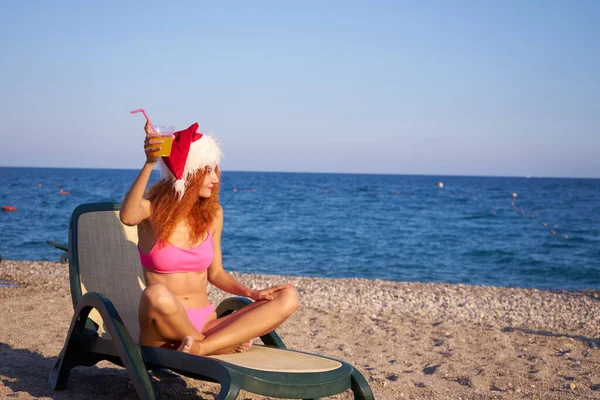 サンタの帽子とピンクのビキニを身に着けている美しい赤い髪の女性は 海の近くのビーチで冬の休暇を楽しんでいます 旅行者の女の子はカクテルを飲む — ストック写真