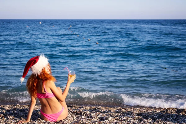 美丽的红头发女人头戴圣诞帽 身穿粉色比基尼 在海边享受寒假 圣诞节或新年假期在海滨的豪华度假胜地 旅行女孩喝燕尾服 — 图库照片
