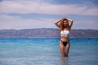 Beyaz kumsalda yürüyen, ince kırmızı saçlı, bikinili güzel bir kızla yaz tatili tarzı. Gezgin kadınlar Salda krater gölü - Burdur, Türkiye 'deki mavi kristal berrak denizde mutlu.                                                       