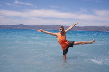 Güneş gözlüklü, yakışıklı, dağların arkasındaki mavi denizde serinleyen genç adamın yaz tatili. Gezgin adamın dinlendiği güzel sahil manzarası.                               