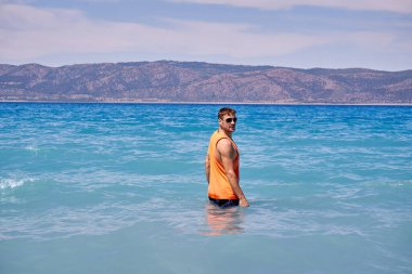 Güneş gözlüklü, yakışıklı, dağların arkasındaki mavi denizde serinleyen genç adamın yaz tatili. Gezgin adamın dinlendiği güzel sahil manzarası.                               