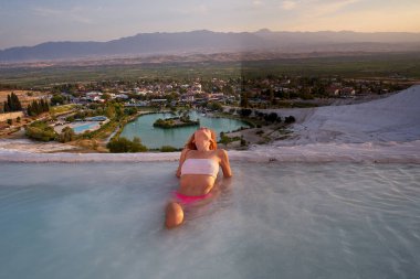 Pamukkale travertine in Turkey with woman enjoying the views. Red hair woman traveler swimming at Pamukkale Natural Park and looking at beautiful sunset.Sexy young woman rest in Pamukkale                                clipart