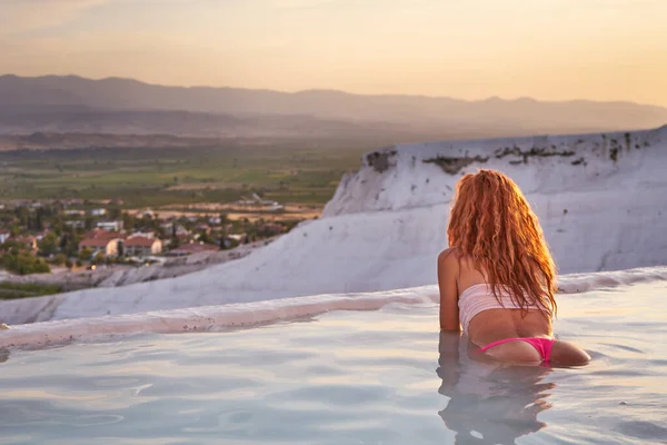 Türkiye 'de Pamukkale traverteni ve manzaranın tadını çıkaran bir kadın. Pamukkale Doğal Parkı 'nda yüzen ve güzel gün batımına bakan kızıl saçlı kadın gezgin. Pamukkale' de dinlenen genç kadın.                               