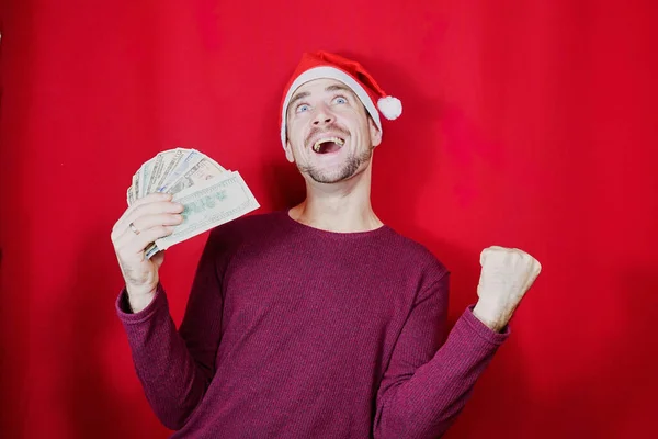 在圣诞节庆祝活动中 戴着圣诞礼帽的快乐的大胡子男人拿着一堆钱 看着镜头和红色背景的人在一起 — 图库照片