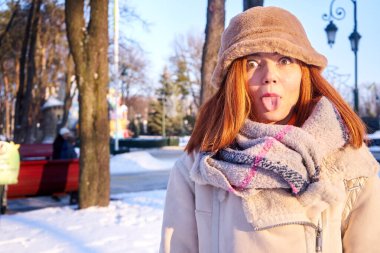 Kar altındaki neşeli kızıl saçlı kız, kışın buzlu vadide dilini gösteriyor. Kış parkında eğlenen, mutlu, genç bir kızın kış portresini kapatıyoruz..                               