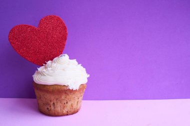 Tasty cupcake decorated with red heart on purple background, space for text. Valentine's Day celebration.Vanilla muffin for romantic date.                                         