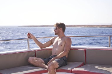 Teknede oturup deniz yolculuğunun tadını çıkaran neşeli bir adamın yat gezisi. Güneş gözlüklü, yatta, mavi deniz koyunda ve gün ışığında gökyüzünde poz veren genç bir adam..                                   