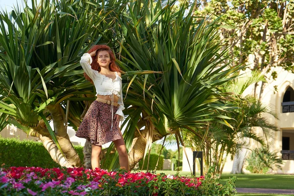 Young beautiful woman stands in the shade of palm trees, view from the back. Red hair girl stays near tropical greenery on a bright sunny day in Egypt.