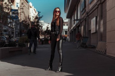 Deri elbiseli, güneş gözlüklü, sokakta poz veren güzel bir kadın.