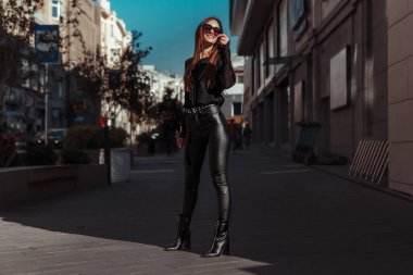 Deri elbiseli, güneş gözlüklü neşeli bayan sokakta gülümsüyor.