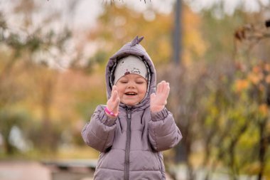 Dinozor ceketli küçük bir kız sonbahar yapraklarıyla oynuyor.