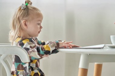 Kafasında at kuyruğu olan şirin bir kız masadaki sandalyede oturmuş dergi okuyor.