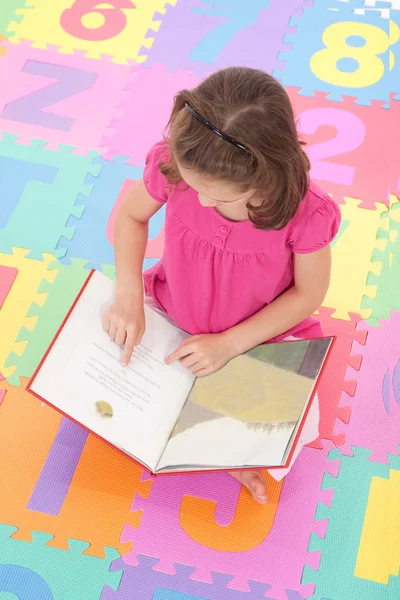 Girl reading book on alphabet floor mat — Stock Photo, Image