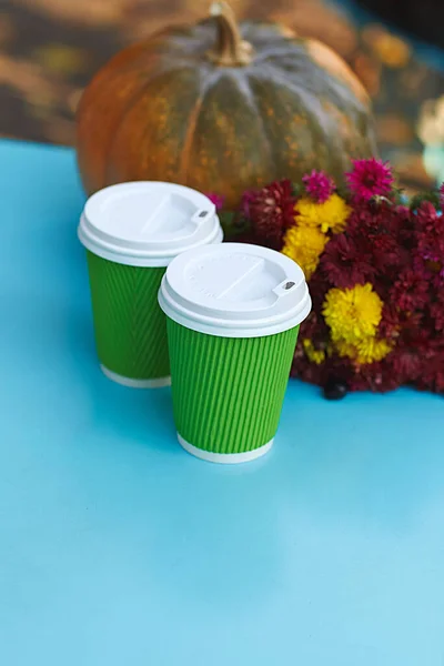 Two green paper disposable cups with coffee or tea on a blue table and autumn background.