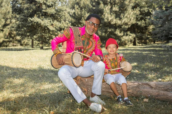 Bello africano in costume nazionale suona un tamburo etnico, djembe insieme al figlio. — Foto Stock