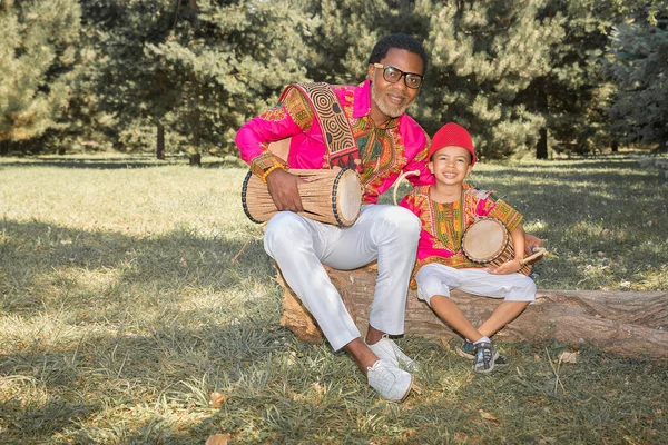 Bello africano in costume nazionale suona un tamburo etnico, djembe insieme al figlio. — Foto Stock