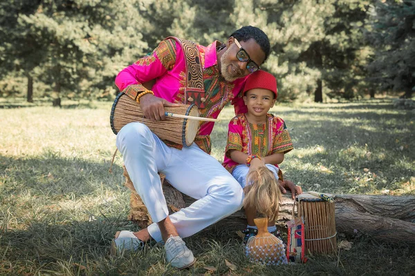 Handsome African man in a national costume plays an ethnic drum, djembe together with son. — Stock Photo, Image