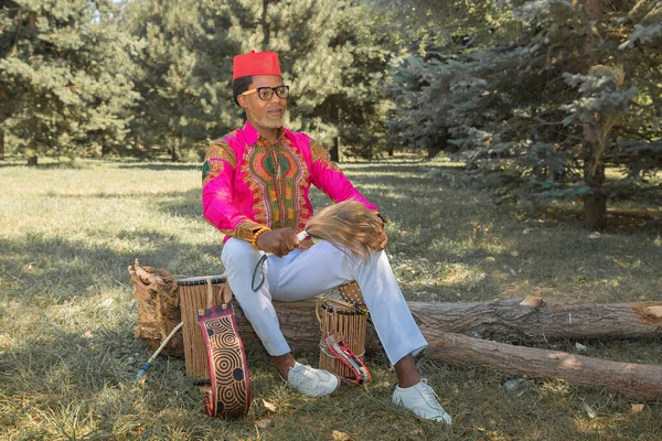 Handsome African man in a national costume plays an ethnic drum, djembe. — Stock Photo, Image