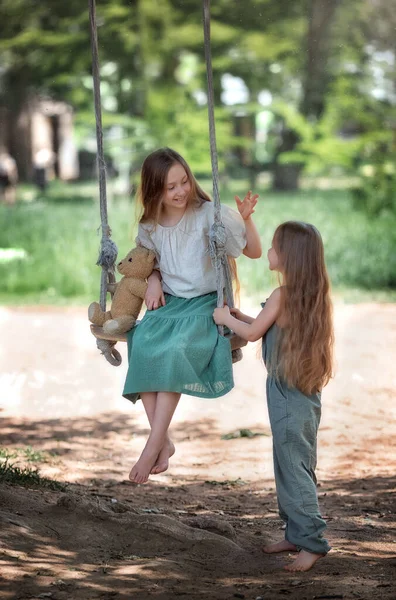夏の晴れた日に公園の遊び場でテディベアのおもちゃでスイングライドを楽しんでいる長い髪を持つ幸せな笑いの子供たちの姉妹 — ストック写真