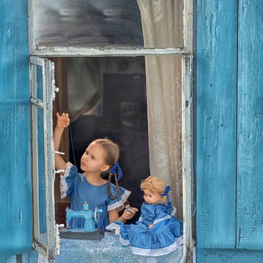 Terzi kızın portresi eski ahşap bir evin penceresinde çocuk dikiş makinesine bebek kıyafeti dikerken görülüyor. Hobi, sanat ve çocukluk konsepti, terzi atölyesi ve terzi