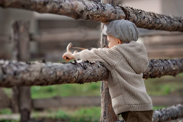 stock image Portrait of bully boy kid with a slingshot aims at someone near a fence in the village outdoors at summer. Rustic barefoot snooty child boy with a hat shoots a slingshot.