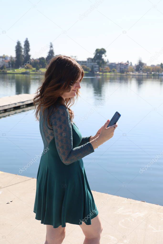 Photo of a Beautiful white girl in a beautiful dress in front of a lake, holding a cell phone